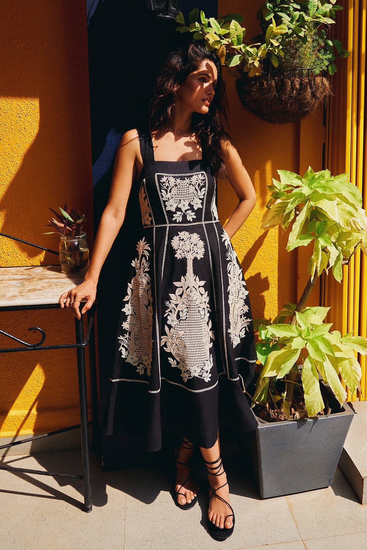 Woman wearing a black dress with white embroidery standing outdoors against a yellow wall with plants.