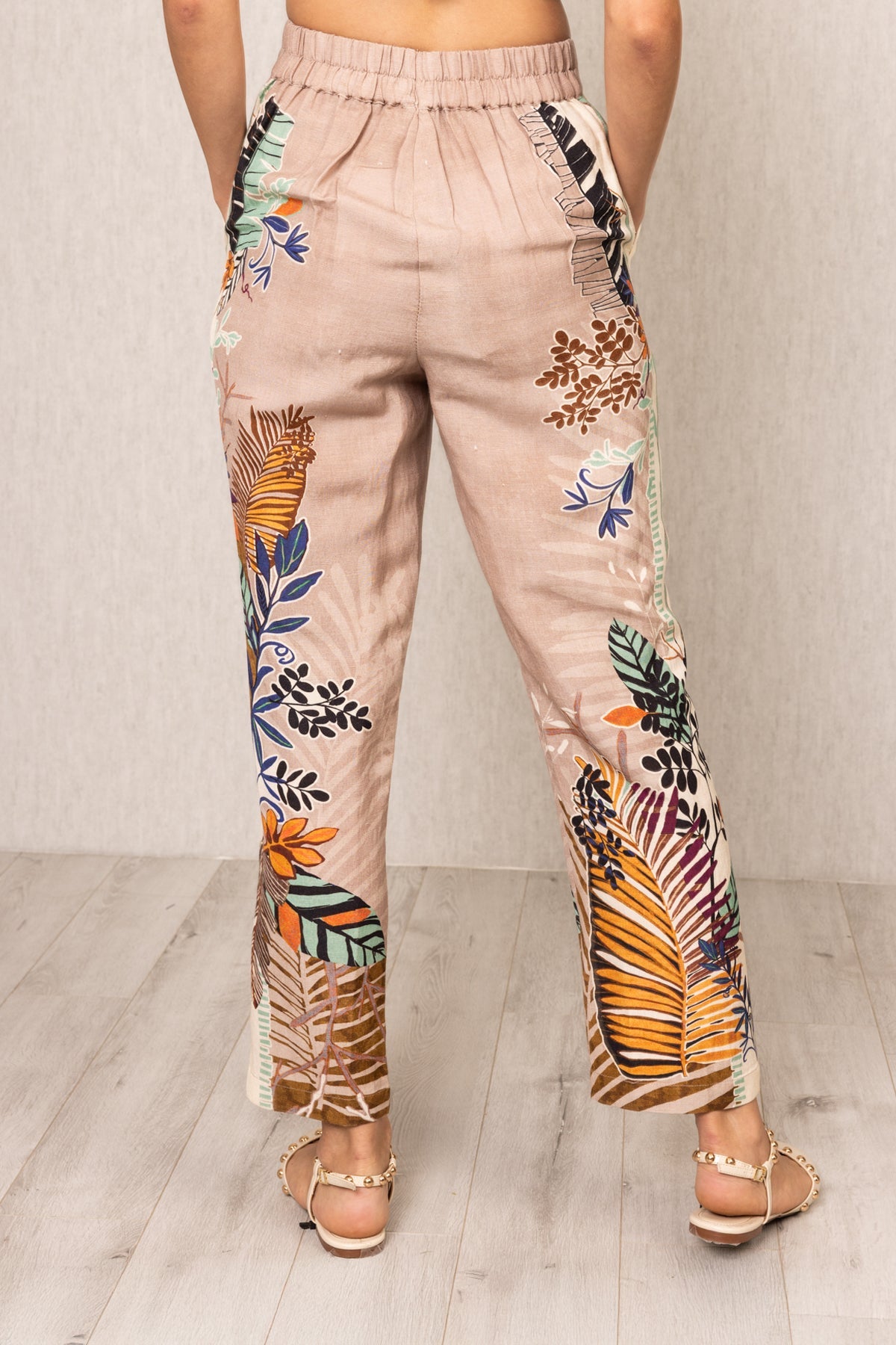 Back view of Ranna Gill Liora Rara Pants in beige linen-viscose fabric with vibrant tropical botanical prints in orange, green, blue, and black, featuring straight fit and elastic waistband.